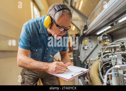Technician with ear protection writing on clipboard in industrial setting Stock Photo
