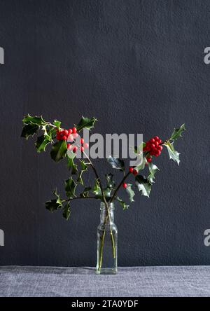 Minimalist festive table decoration of cut holly branches with red berries and leaves, in a reused condiment bottle, on a grey background. Stock Photo