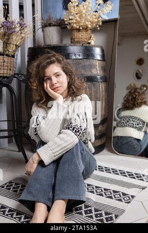 young woman, beautiful teenage girl of 17 years old in a sweater and jeans sits dreamily on the floor in a cozy room. rest, harmony of life, self-refl Stock Photo