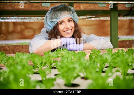 Happy woman agronomist checking plant growth in greenhouse. Joyful female gardener looking at green leafy plants and smiling while standing near shelf with seedlings. Stock Photo