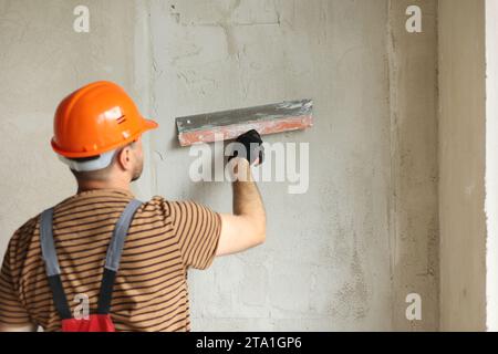 Rear view of handyman worker in overalls and protective helmet plastering concrete wall with putty using a big putty knife. Renovation at apartment an Stock Photo