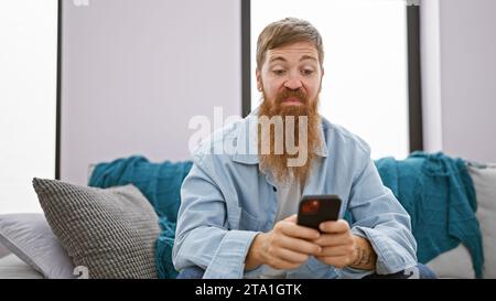 Focused young redhead man, chilling on his sofa indoors, engrossed in texting on his smartphone at his cozy apartment. Stock Photo
