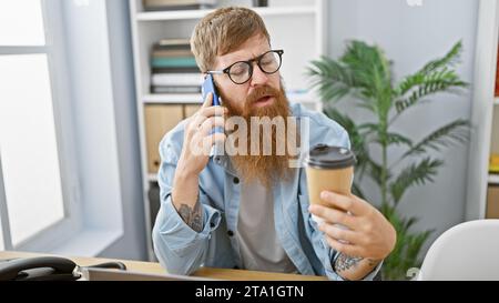 Intriguing sight of a young, handsome, redhead business man engrossed in serious talk on his smartphone, drinking espresso coffee while at the office, Stock Photo