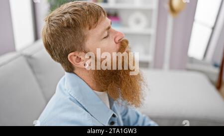 Handsome young redhead man sitting alone on living room sofa indoors, breathing deeply with closed eyes, displaying serious expression, while wrestlin Stock Photo