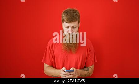Focused redhead man engrossed texting on smartphone, serious face over isolated red background Stock Photo