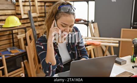 Charming young hispanic woman carpenter juggling business with craftsmanship, engrossed in online conversation on smartphone amid sawdust at her indoo Stock Photo