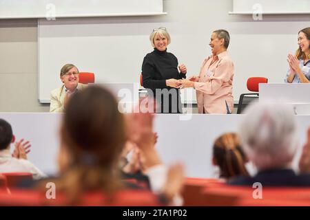 Female orators handshaking on stage in front of audience at business conference event Stock Photo