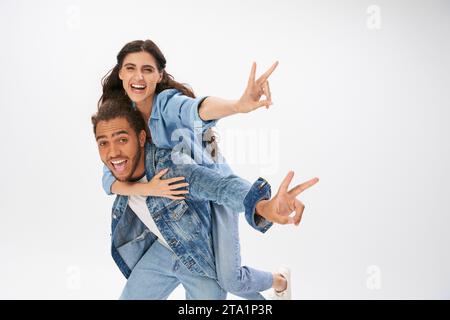 overjoyed couple in trendy denim wear showing victory signs while having fun on grey, piggybacking Stock Photo
