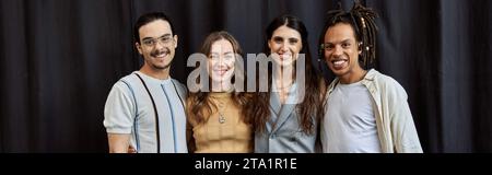 cheerful multiethnic startup team in casual attire standing near black drape in office, banner Stock Photo