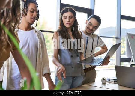 female team lead with laptop pointing with finger during meeting with multiethnic team in office Stock Photo