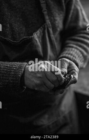 Monochrome photo of hands of Senior violinmaker wearing apron holding a tool chisel in workshop in Cremona, Italy Stock Photo