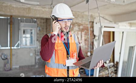 Young hispanic man builder talking on walkie talkie using laptop at construction site Stock Photo