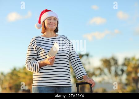 Independent mature woman in Christmas Santa Claus hat, striped shirt holds passport, boarding ticket, suitcase on seashore with palms backdrop celebra Stock Photo