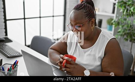 Delightful african american business woman, confidently working online with laptop, drinking coffee at office desk. Stock Photo