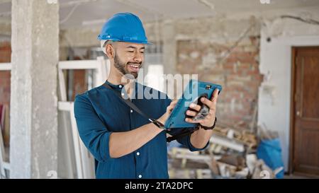 Young hispanic man architect wearing hardhat smiling using tablet at construction site Stock Photo