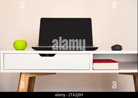 Empty open laptop with mockup and black screen on white desk table. Green apple and mouse. Book. Modern workspace Stock Photo