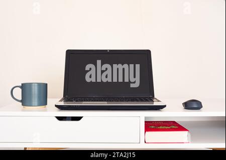Empty open laptop with mockup and black screen on white desk table. Coffee mug and mouse. Book. Modern workspace Stock Photo