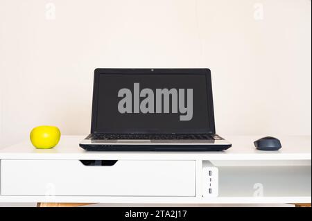 Empty open laptop with mockup and black screen on white desk table. Yellow apple and mouse. Charger. Modern workspace Stock Photo