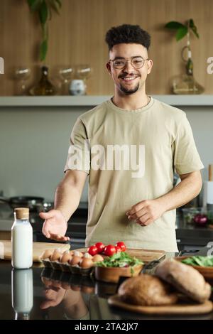 Medium shot of smiling young middle eastern guy showing food on kitchen table Stock Photo
