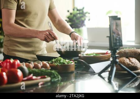 Medium shot of unrecognizable man standing by stove at kitchen frying food in pan while being filmed on smartphone Stock Photo