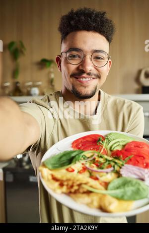 Headshot of smiling young middle eastern guy taking picture of himself holding plate with omelet and vegetables Stock Photo