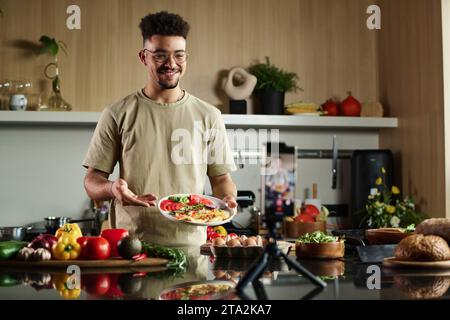 Medium shot of young middle eastern influencer demonstrating food to smartphone camera while making video for blog Stock Photo