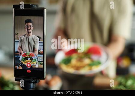Closeup of mobile phone on tripod filming young middle eastern blogger showing plate with omelet and vegetables, focus on smartphone, copy space Stock Photo