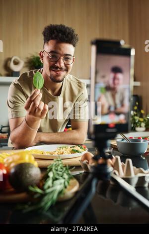 Waist up portrait of smiling young middle eastern blogger at kitchen showing leaf of spinach to smartphone camera making video for his channel Stock Photo