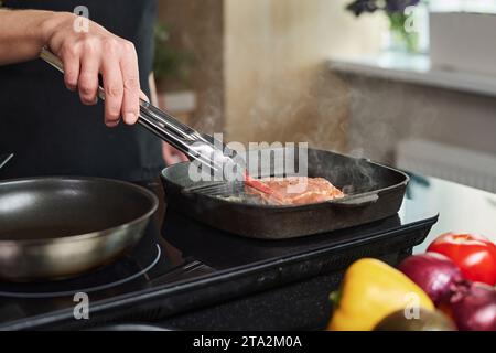 Closeup of hand of unrecognizable man placing chili in pan near cooking piece of meat using metal tongs Stock Photo