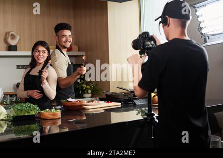 Middle eastern man and hispanic woman standing back to back holding forks with food by kitchen counter while being photographed by cameraman Stock Photo