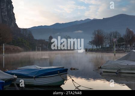 Lake Lugano at the strait of Lavena Ponte Tresa at sunrise, mist on the water and moored boats in the foreground Stock Photo