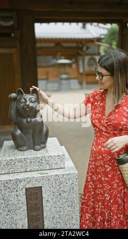 Beautiful hispanic woman in glasses counting on luck, standing by the famous lucky cat statue at gotokuji temple in tokyo Stock Photo