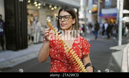 Beautiful hispanic woman joyfully eating crunchy, delicious chips on a stick at takeshita street, tokyo - traveling junk food lover, sporting glasses Stock Photo