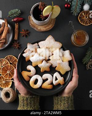 Cookies in the form of numbers 2024 and stars on the dark plate holding by hands and dark background, decorated with pine twig Stock Photo