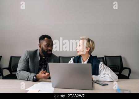 Focused middle-aged female leader helping new black worker with computer corporate software application in office. Motivated diverse employees Stock Photo