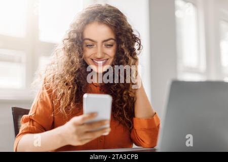 Happy curly-haired woman using smartphone at workplace Stock Photo