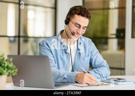 Smiling handsome young european man manager in casual with laptop at table with headphones taking notes Stock Photo