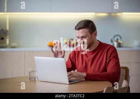 Happy glad man has online meeting video call sits on kitchen waving hand looking at laptop screen. Stock Photo