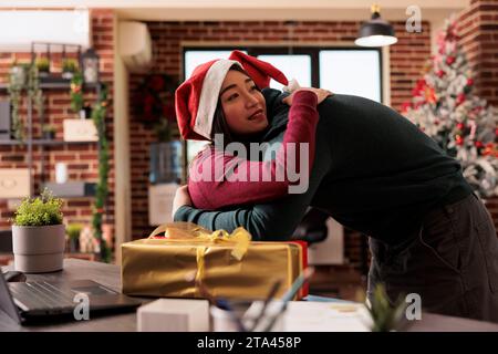 Diverse colleagues hugging while sharing christmas gifts in startup company decorated office. Man and woman coworkers embracing while working and exchanging presents at xmas eve Stock Photo