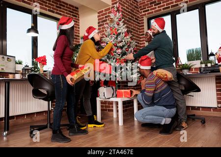 Diverse colleagues sharing xmas presents at winter seasonal holiday party in decorated office. Happy coworkers in santa hats exchanging gifts near christmas tree with ornaments Stock Photo
