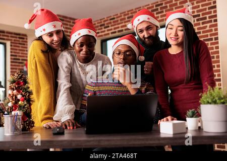 Cheerful colleagues checking sales annual report results on laptop before celebrating christmas in office. Smiling happy coworkers working for project deadline together in festive decorated workplace Stock Photo