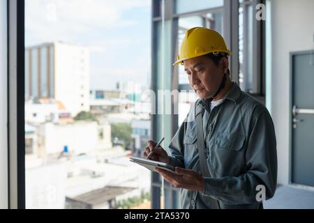 Contractor wearing hardhat when working at construction site, filling form on tablet computer Stock Photo