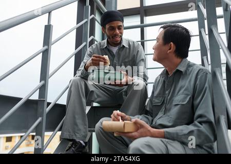 Positive construction worker eating sandwich and talking to coworker during lunch break Stock Photo
