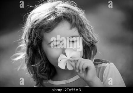 Funny child smelling plumeria flower, face close up. Kids in summer nature park, portrait. Stock Photo