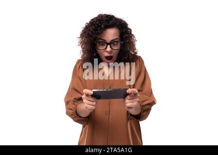 young curly brunette woman dressed in a brown shirt plays a game on a mobile phone Stock Photo