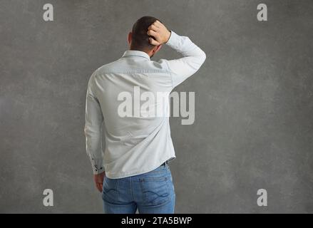 Unknown man stands with his back to the camera and scratches the back of his head in confusion. Stock Photo
