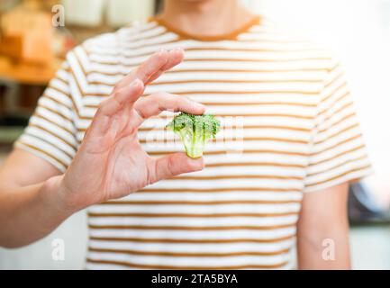 Front view and selective focus of man in striped t shirt holding broccoli piece showing to camera. Sunny day inside the kitchen Stock Photo