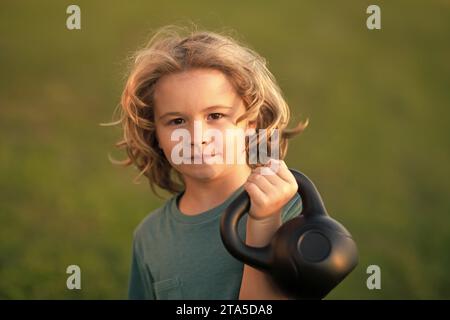 Child lifting the kettlebell in park outside. Child boy pumping up biceps muscles with kettlebell. Fitness kids with dumbbells Stock Photo