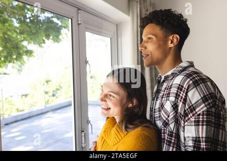 Happy diverse couple embracing and looking through window at home, copy space. Expression, leisure, love, togetherness and domestic life unaltered. Stock Photo