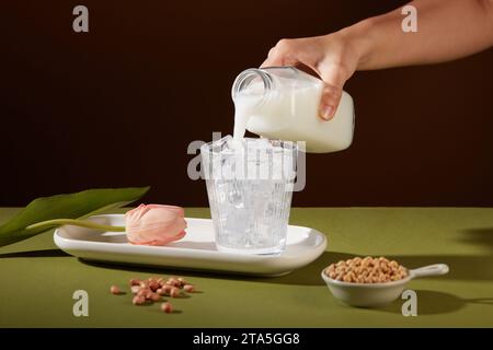 A hand is pouring milk from a bottle into a cup with ices inside. Soybeans contained in a bowl and some peanuts displayed. Nut milk has many advantage Stock Photo